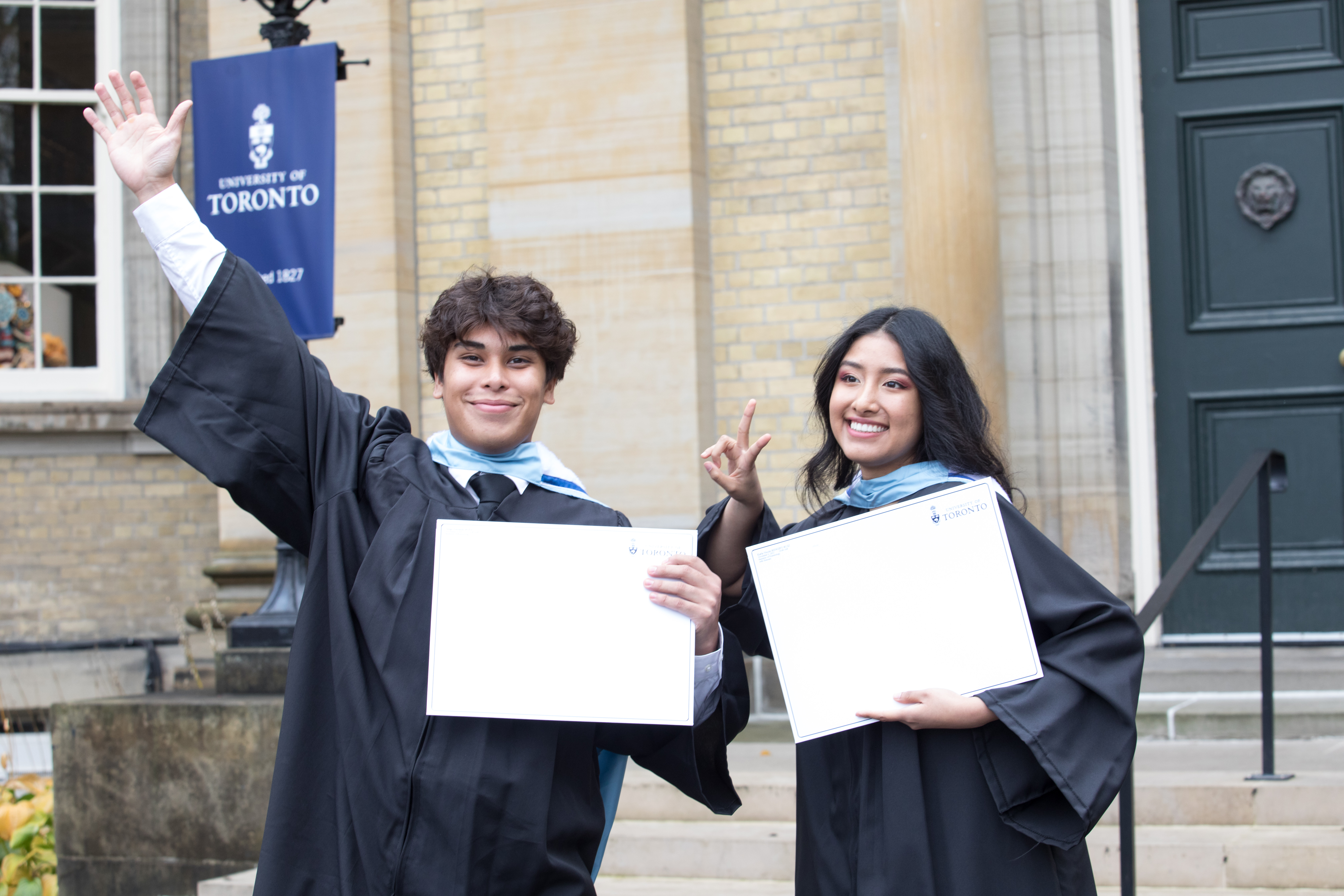 two smiling students outside of simcoe hall in graduation gowns, holding envelope with degree