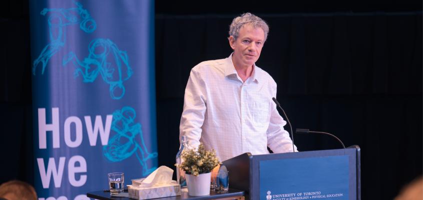 Dr. Ian Ritchie stands behind a KPE faculty podium on stage at Innis College. Behind him is a How We Move banner, and on his table is a potted plant and a box of tissues.