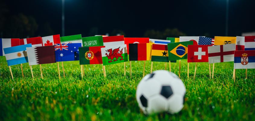 An out-of-focus soccer ball sits in the foreground on a green grass field in front of a number of small flags denoting the nations participating in the FIFA World Cup.