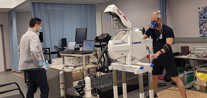 A researcher stands in front of a computer console while a study participant uses an arm bike while wearing an array of testing equipment attached to his face.
