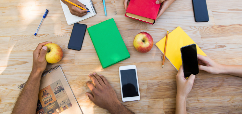 overhead shot of desk with arms in frame, surrounded by notebooks, apples and cellphones