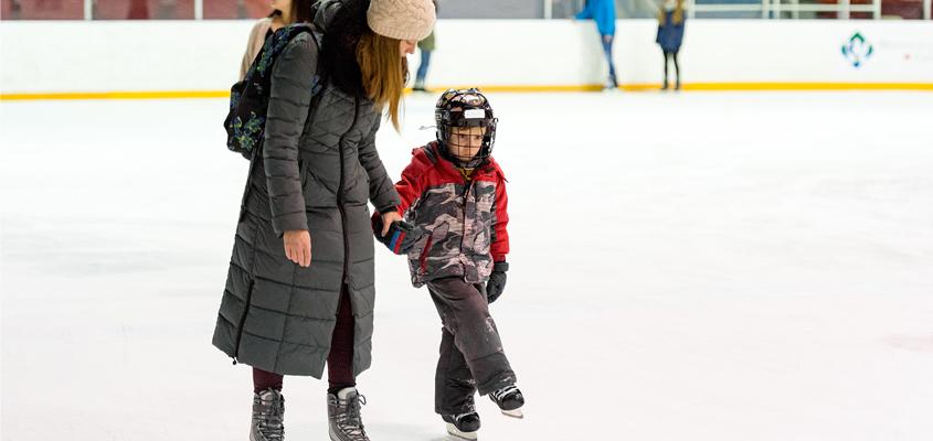 mother holds child's hand as they ice skate at rink