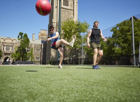 two young males in shorts on a sunny day kicking a ball on a turf field