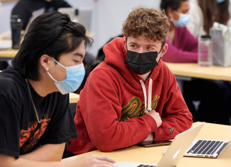 two male students wearing medical masks chatting in class in front of laptops